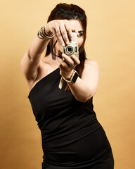 Studio portrait of a stylish woman in a black dress holding a camera toward the viewer against a golden background. Symbolizes creativity, empowerment, and confidence in modern fashion.