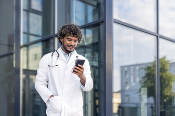 Smiling Indian man doctor standing in white uniform on the street and using mobile phone