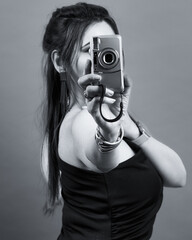 Black and white studio portrait of a stylish woman in a black dress holding a camera toward the viewer, representing confidence, creativity, and empowerment in a minimalist, modern composition.