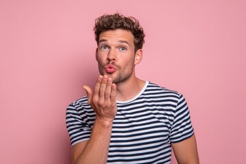 Attractive young man in a striped tee against a pink backdrop blowing a kiss and radiating charm