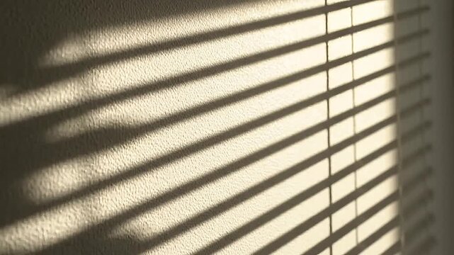 Warm sunlight streams through venetian blinds, casting dramatic shadows on a textured wall.