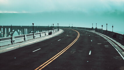 Fototapeta premium A quiet road bends gently beside a calm lake, surrounded by misty skies. Street lamps line the way, enhancing the peaceful atmosphere of this tranquil waterfront scene.