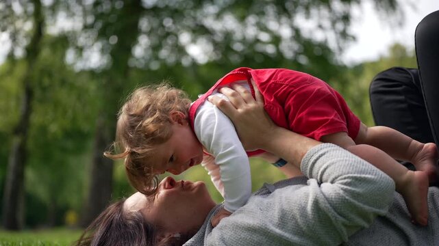 Mother holding baby up over her face in joyful play on sunny day, both smiling and connecting affectionately during happy bonding moment in lush green park