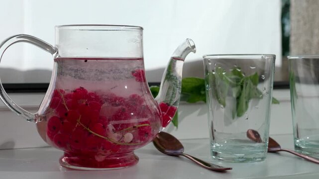Still life: a glass teapot with red currant tea is steeping as sediments settle at the bottom. Summer mood and mint in the background.