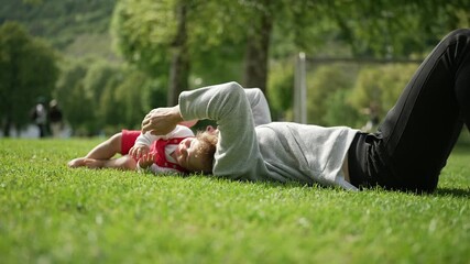 Mother lying on grass with toddler beside her in open park, sharing tender bonding moment surrounded by trees and sunlight, symbolizing love, connection, and family warmth outdoors