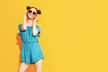 Young beautiful smiling female in trendy summer jeans overalls. Sexy carefree woman with two horns hairstyle posing near yellow wall in studio. Positive model having fun. Isolated. In sunglasses