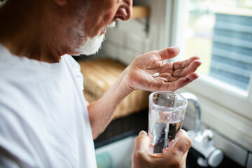 Senior man taking medicine with water in home kitchen, focused