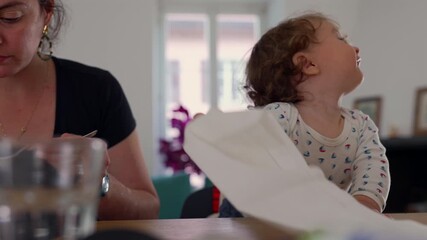 Toddler being fed with spoon by parent at dining table while playing with napkin, showing gentle interaction and curious behavior during relaxed family mealtime