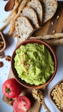 Wooden bowl filled with creamy avocado guacamole dip, garnished with pine nuts and served on a board with sliced whole grain bread, breadsticks, and fresh tomatoes for a healthy appetizer