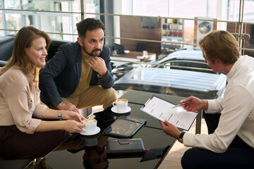 Caucasian young adult woman and man sitting at table discussing contract with Caucasian young adult man holding clipboard in modern car dealership office