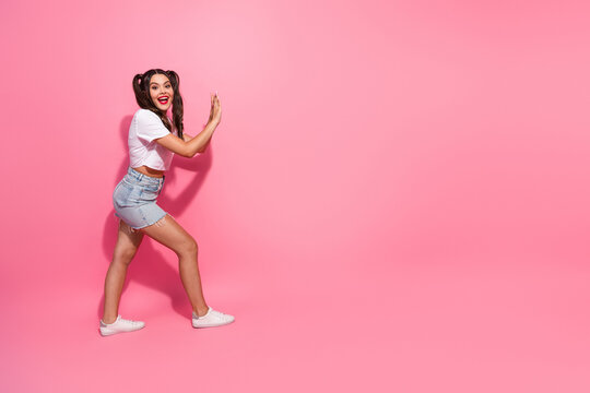 Young woman in casual white top and denim shorts smiles broadly as she playfully poses against a bright pink studio background