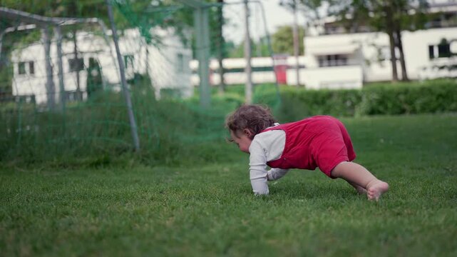 Toddler crawling on green grass near soccer goal net in city park, showing curiosity and energy in playful outdoor setting surrounded by buildings and trees