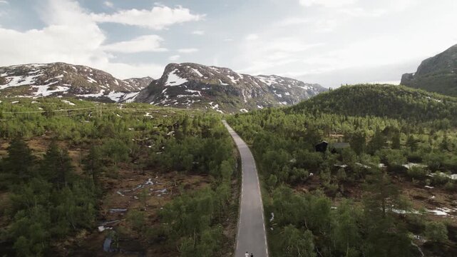 Aerial view of a straight road cutting through a dense green forest, leading toward majestic mountains with patches of snow, Dalen, Telemark, Norway.