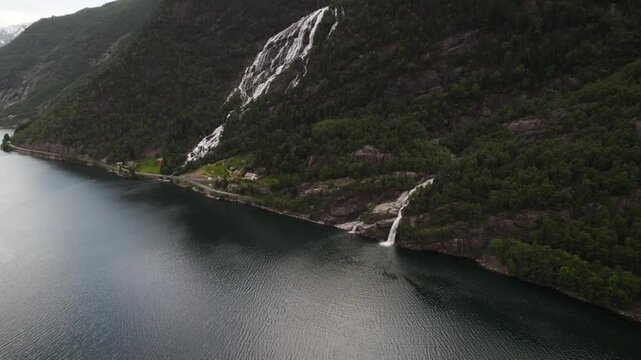 Aerial view of cascading waterfalls flowing down rocky mountains covered in lush green trees, creating a beautiful contrast with the dark water, Odda, Vestland, Norway.