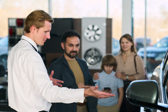 Caucasian middle aged man gesturing while explaining car features to Middle Eastern middle aged man with beard and Caucasian woman and child - Powered by Adobe