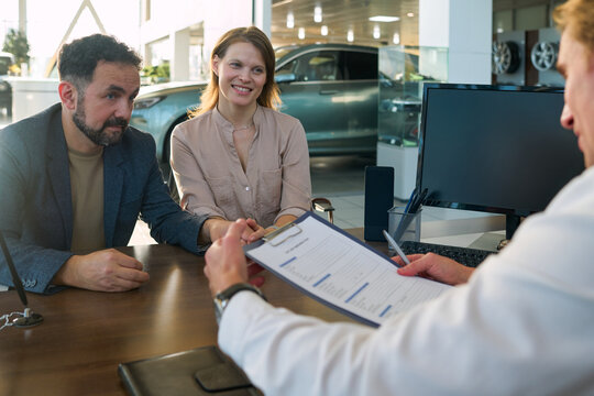 Caucasian young adult man and Caucasian young adult woman sitting together at desk listening to middle aged Caucasian man holding clipboard discussing car dealership paperwork