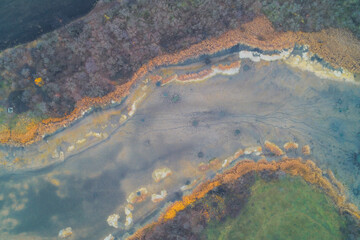 Abstract drone shot capturing dry riverbed or marsh area. The center is dominated by cracked, gray soil with circular depressions, bordered by dried, colorful reeds: yellow, orange and dark vegetation