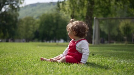 Toddler sitting on grass in open park, looking away toward trees and distant soccer goal, evoking calm reflection and peaceful connection with nature in early childhood