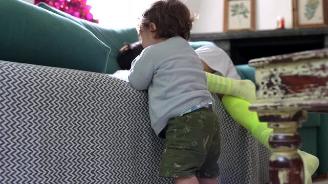 Toddler standing barefoot near sofa touching edge with cautious curiosity while learning to balance, surrounded by scattered toys on living room floor in cozy home environment