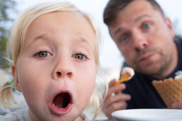 Dad and Little Girl Sitting in a Cafe, Eating Ice Cream and Looking Surprised at the Camera