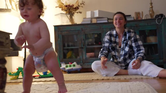 Toddler taking first independent steps while trying to keep balance, stumbling but determined to stand again as mother encourages with pride and joy in background