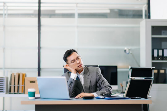 Exhausted businessman sitting at desk with laptop, stressed by workload, deadlines, and corporate pressure in modern office environment. - Powered by Adobe