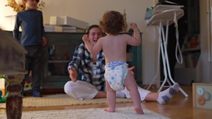 Toddler taking first steps toward mother in living room, stumbling and standing again with determination as mother cheers joyfully, capturing authentic family milestone and emotional moment