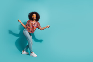 Young woman with Afro wearing patterned shirt jeans and white sneakers posing playfully in blue...
