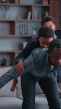 Carefree African American family playing piggyback pretending airplane at home joyful father dad riding on back little son play plane flying pretend with two kids boys sons children having fun joy