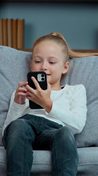 Caucasian active little child daughter kid girl jump on sofa using mobile phone apps gadget addict generation falling on couch in living room scrolling smartphone social media networks internet chat