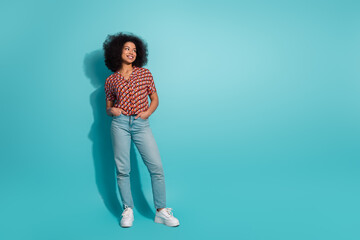 Young woman with curly hair wearing a red printed blouse and light blue jeans posing against a bright turquoise studio background