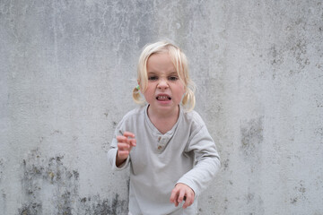 Little 4 Year Old Blonde Girl Standing by a Wall, Pretending to Be Angry and Showing Claws