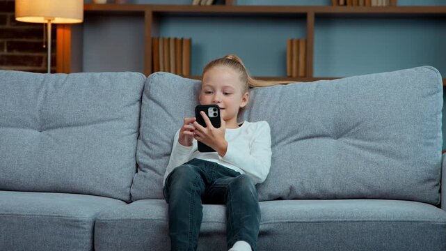 Caucasian active little child daughter kid girl jump on sofa using mobile phone apps gadget addict generation falling on couch in living room scrolling smartphone social media networks internet chat