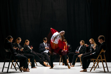 Expressive leap of performer in red jacket with motion-blur amid colleagues absorbed in smartphones. Concept of artistic contrast, creative individuality, emotional expression and stage identity.
