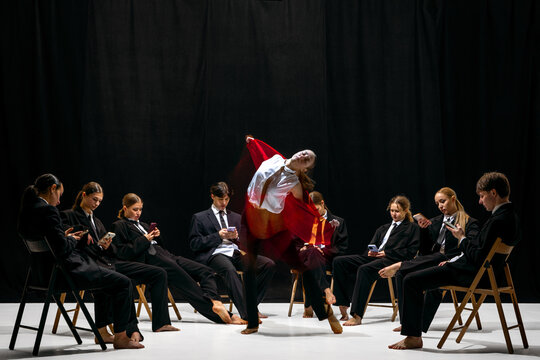 Expressive leap of performer in red jacket with motion-blur amid colleagues absorbed in smartphones. Concept of artistic contrast, creative individuality, emotional expression and stage identity.
