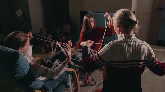Mother with her two teenage daughters untangling yarn together by the fireplace, laughing and enjoying a cozy winter evening filled with handmade crafts and family warmth.