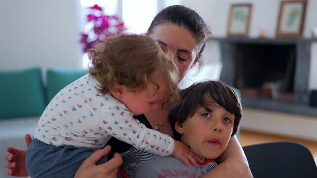 Mother holding toddler and older child as toddler pushes sibling away, wanting exclusive attention and creating emotional family moment at home