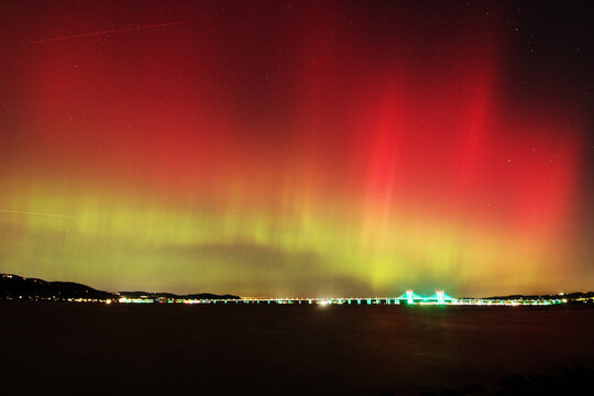 View of the crimson and emerald aurora borealis dances above the glittering lights of the bridge, a celestial ballet over the dark water, Piermont, New York, United States.