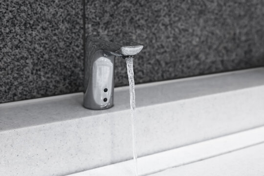 A close-up, black and white shot of an automatic sensor faucet running with a stream of water into a white basin.