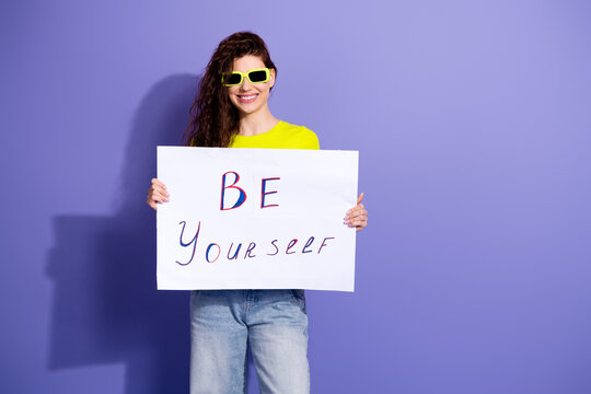 Young woman in neon shirt holds a sign be yourself against purple background for bold inspirational lifestyle and fashion stock content
