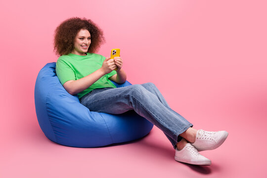Young woman relaxes on a blue bean bag uses yellow smartphone against pink background casual style - Powered by Adobe