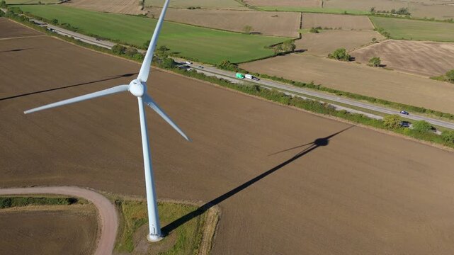 Aerial view of a towering wind turbine casting a long shadow over the brown fields, near a highway with passing vehicles, Melton Mowbray, England, United Kingdom.