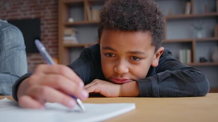 Frustrated boring African American boy son writing homework sad kid child pupil fatigued with primary elementary education studying at home lying on table bored learning knowledge difficulty stressed - Powered by Adobe