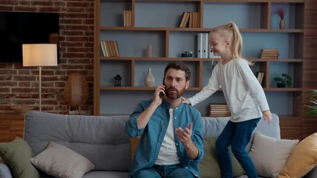 Caucasian family at home father talking phone naughty daughter jumping on couch angry dad businessman talk mobile call distant negotiate annoyed with active little kid child girl distract jump on sofa