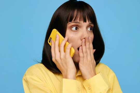 A surprised young woman talks on a bright yellow cellphone against a vivid blue background, covering her mouth with her hand and widening her eyes in astonishment.