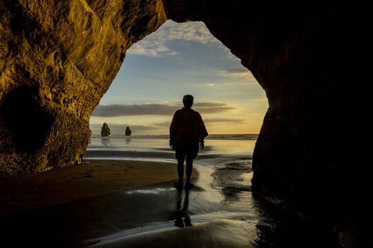 View of a silhouetted figure walking on the beach through a natural archway towards distant rock formations under a soft, golden sky, Tongaporutu, Taranaki Region, New Zealand.