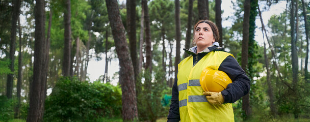Young confident female lumberjack wearing work clothes and reflective vest holds a yellow protective helmet under her arm in the forest