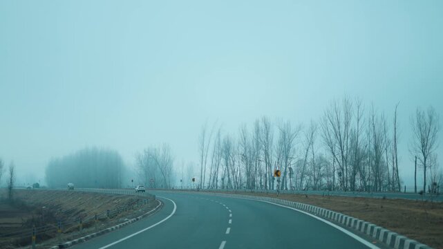 Morning fog by the road with leafless tree branches on Highway at highway in Srinagar, Kashmir, India. POV shot of car driving on dreamy mountain road. Roadtrip and travel concept.