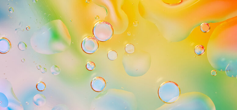 Macro photograph of water droplets against a bright background with smooth transitions of yellow, green, orange, and blue colors. texture is fluid, with bubbles of various sizes, light accents. Banner