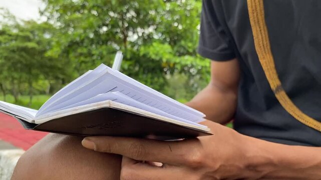 asian man sitting outdoors, holding a notebook, and writing with a pen. Ideal for themes of creativity, study, leisure, mental health, and travel journaling.